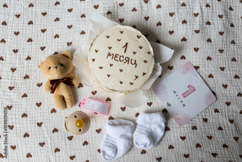 Birthday cake for a newborn, with baby accessories and elements for a baby milestone photo shoot. Children's socks, a pacifier, a soft toy and a card with the number one are neatly laid out in the