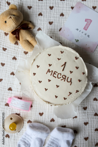 Birthday cake for a newborn, with baby accessories and elements for a baby milestone photo shoot. Children's socks, a pacifier, a soft toy and a card with the number one are neatly laid out in the