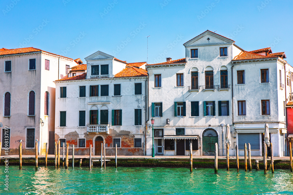 Obraz premium Historic buildings line the Grand Canal in Venice under a blue sky. Wooden posts stand in the vibrant turquoise water in the foreground