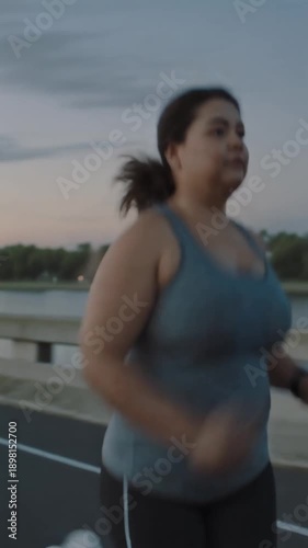 Determined plus-size woman jogging outdoors on a bridge during a sunset or sunrise workout session.