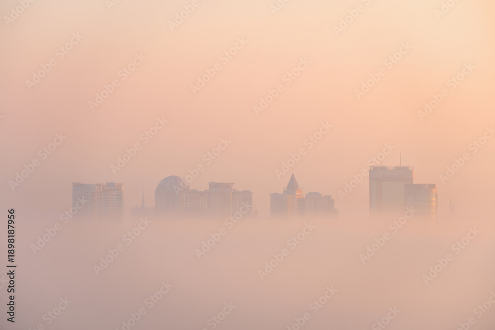 Obraz premium Ethereal cityscape shrouded in dense, atmospheric fog at sunrise or sunset, with prominent buildings softly emerging from the warm, peachy-orange mist. A tranquil and mysterious urban scene.