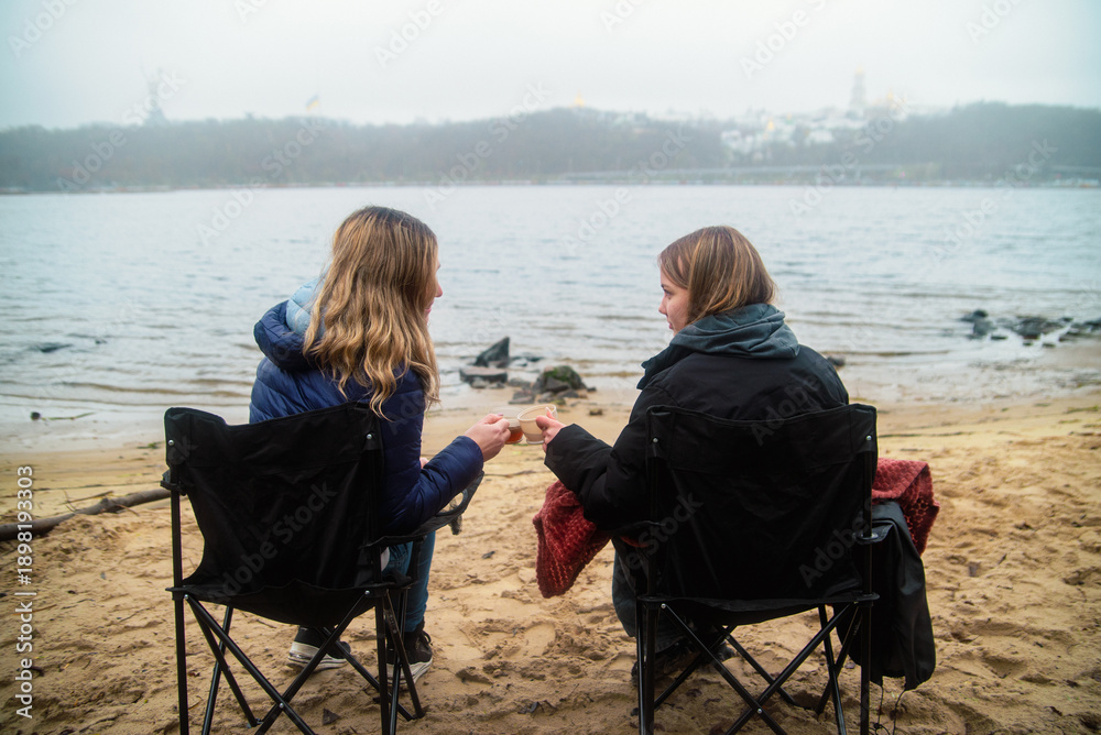 Fototapeta premium Two women sitting in folding chairs on a sandy beach, drinking hot beverages on a foggy day in Kyiv