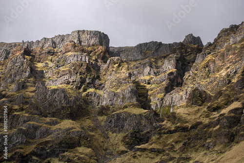 A dramatic view of a rugged mountain cliff covered with patches of moss and grass under a cloudy sky. This natural landscape captures the raw and untamed beauty of highland terrain. Iceland
