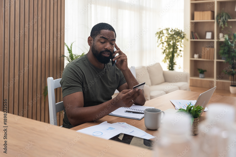 Fototapeta premium Male investor at home buying gold shares and funding AI infrastructure, smiling on phone while paying with credit card, reviewing market charts on laptop in modern living room.