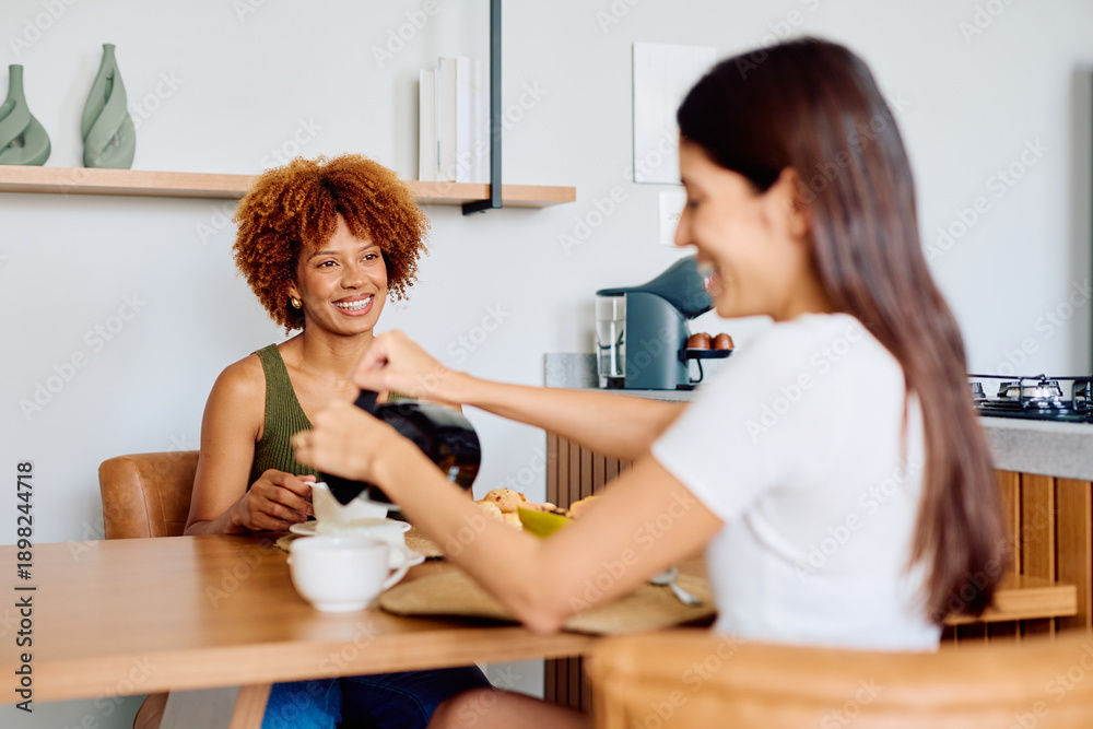 Obraz premium Two cheerful young women enjoying a relaxed breakfast together, laughing while pouring coffee at a kitchen table