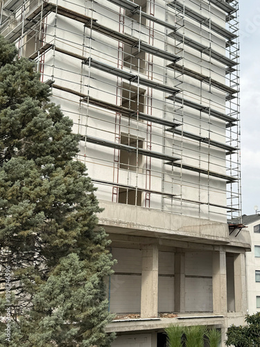 Urban apartment building under construction with full scaffolding and unfinished facade. Vertical view with tree foreground, showing renovation progress, architecture and real estate development.