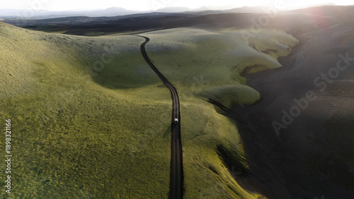 Aerial view of a lone vehicle winding along a dark path through rolling green hills under the soft glow of the setting sun, Selfoss, Iceland.