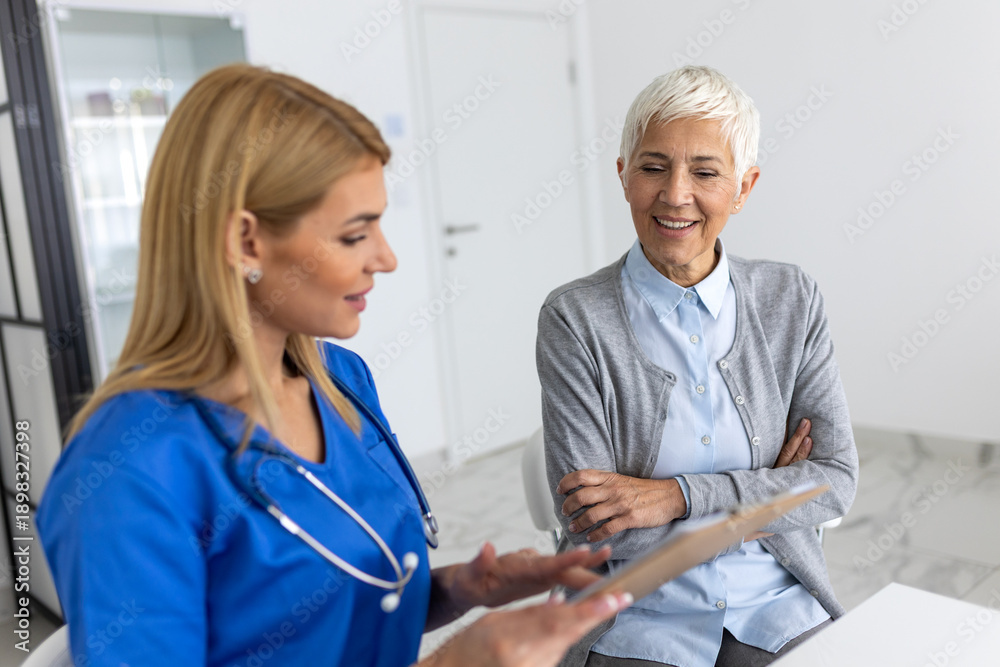 Fototapeta premium Professional female doctor in blue scrubs discussing medical results on a clipboard with a smiling senior woman during a private consultation in a modern healthcare clinic.