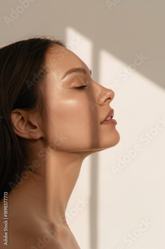 Side profile portrait of a woman with closed eyes and glowing natural skin illuminated by soft sunlight creating geometric shadows on a neutral background 