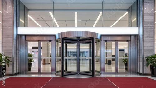 Entrance to modern office or commercial building with central revolving door and automatic glass doors. Red carpet, stainless steel, glass panels and plants lead to bright lobby
