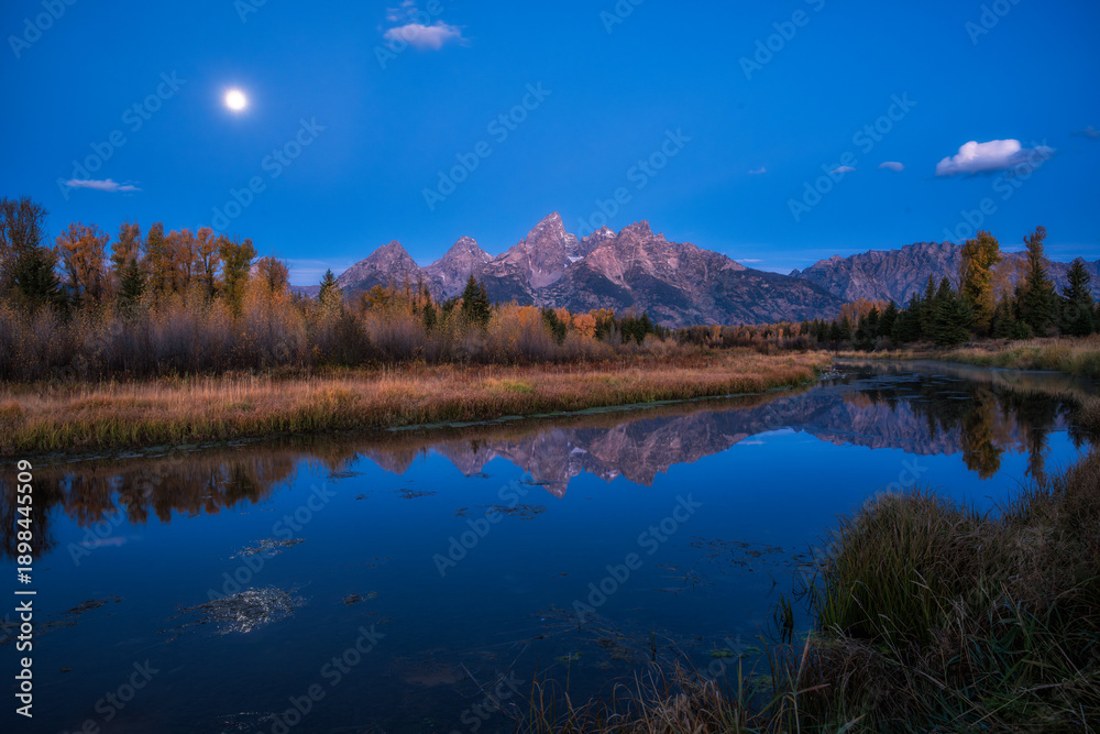 Fototapeta premium A high-resolution landscape of the Teton Range reflected in a calm river during twilight, featuring vibrant autumn foliage and a bright moon in a deep blue sky.