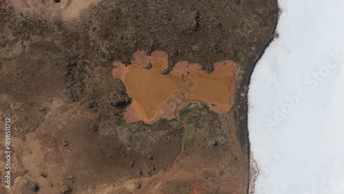 Aerial view of a rusty-red pond nestled amidst the rugged terrain, contrasting sharply with the stark white shoreline, Selfoss, Iceland.
