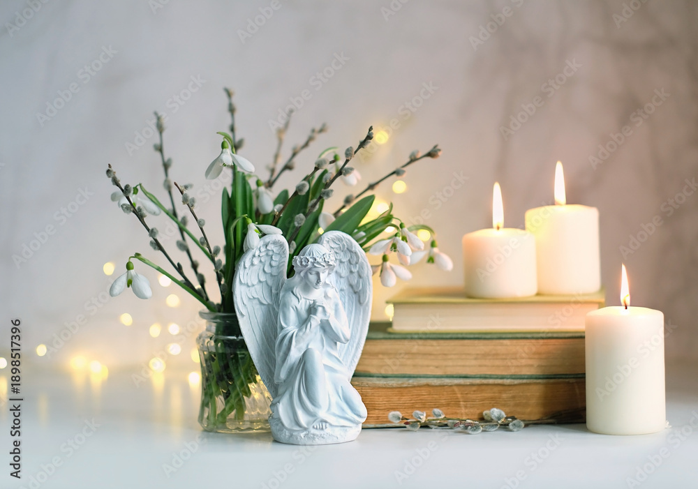 Naklejka premium praying angel figurine, burning candles, books, snowdrop flowers on table, abstract light background. Christmas or Easter holiday concept. Christian Religious church fest. symbol of faith in God