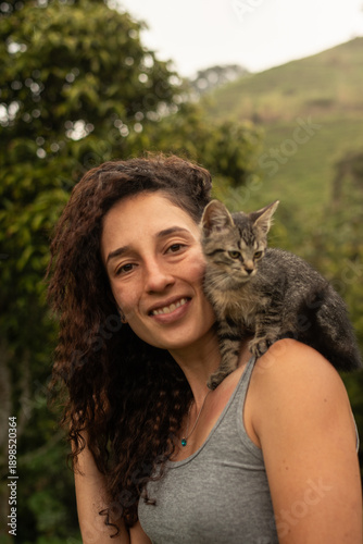 Happy Hispanic woman smiling with teeth visible, tilting her head affectionately towards a small tabby kitten perched on her shoulder. Outdoor rural setting with green mountains in the background.