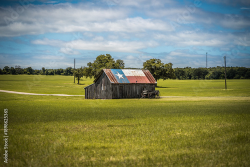 Old Rustic Barn With Texas Flag Painted on Roof With Old Wagon Nearby In Green Field in Rural Town in Texas