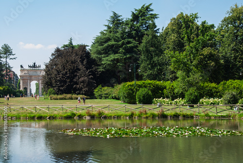 Peaceful view of Parco Sempione in Milan with a reflective pond, lush greenery and the Arco della Pace in the background on a sunny summer day.