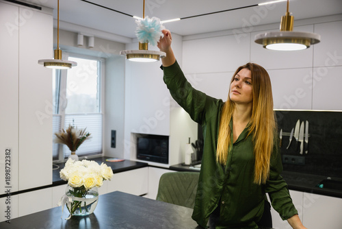 Woman wiping dust from a hanging lamp in a modern kitchen interior. Home cleaning routine, everyday household activity, minimalistic apartment, natural light.