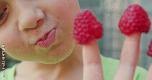 Young boy opens mouth biting raspberry covering fingertip. Child chews berry licking juice while hand withdraws to grab next fruit during fresh snack time