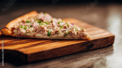 Slice of tuna pizza topped with green peppers and onions on a wooden cutting board, with a blurred background highlighting the food presentation