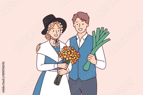A young couple dressed in authentic Welsh national costumes smiles while holding traditional symbols like yellow daffodils and a large green leek to celebrate Saint David's Day in Wales.