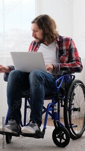 A young disabled man in a wheelchair works on his laptop with documents at home. Vertical video.