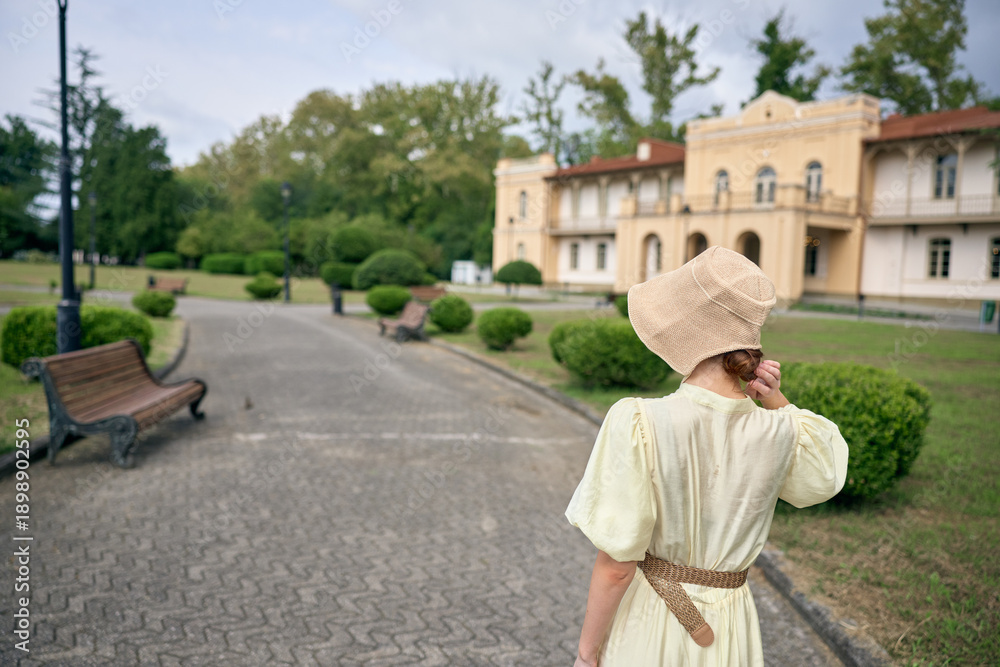 Fototapeta premium Woman in a vintage dress and straw hat walks along a park path toward a grand estate, creating a serene outdoor scene with timeless charm today.
