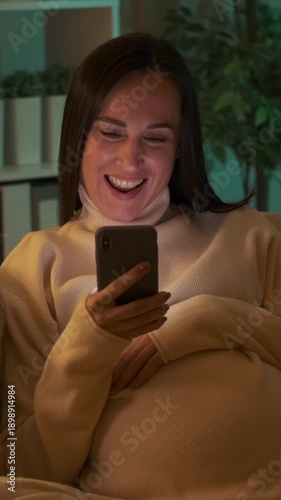 A happy pregnant woman uses her smartphone and touches her belly while relaxing on the sofa at night. Vertical video.