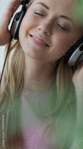 Vertical video of a happy young blonde woman with headphones on, closing her eyes and smiling while enjoying music at home.