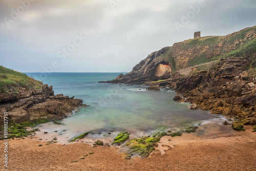 View of Santa Justa beach, with the Santa Justa Hermitage integrated into the rock, near Santillana del Mar, in Cantabria, Spain, on a cloudy summer day.