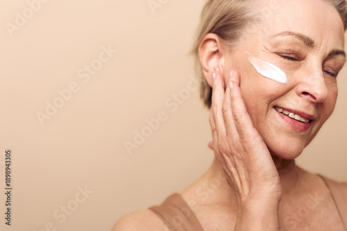 Tableau sur toile Smiling mature woman applies moisturizing cream on her cheek with closed eyes, enjoying skincare routine