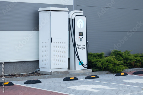 Electric vehicle fast charger station with charging unit and power supply cabinet located in a parking area beside green shrubs and gray building wall
