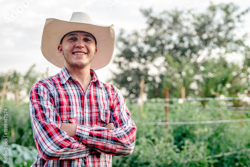 Vászonkép Portrait of a smiling young male farmer wearing a cowboy hat and red plaid shirt standing with arms crossed in a rural field