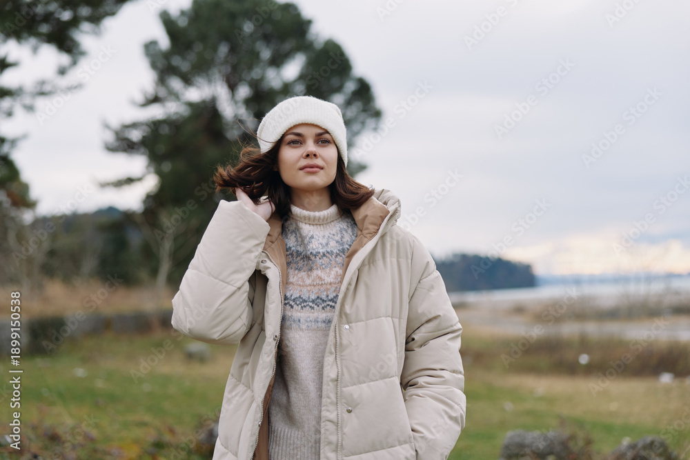 Fototapeta premium Winter woman outdoor fashion coat portrait in chilly countryside with sweater and beanie, soft light, calm landscape, cozy look for cold weather lifestyle.