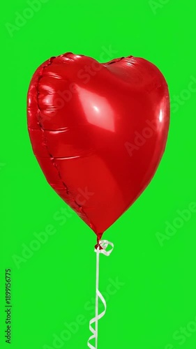 A red heart-shaped balloon floats gently with a white string against a vibrant green background as seen from a straight viewpoint