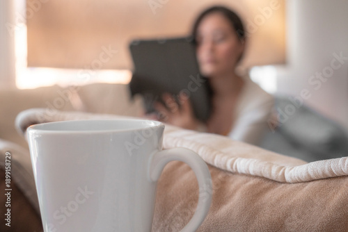Fotografia Primer plano de taza de café blanca con fondo desenfocado de mujer, viendo tableta digital en la sala de estar
