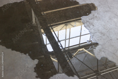 A puddle of fuel and water on a concrete floor with reflections of the ceiling and window structures inside the building