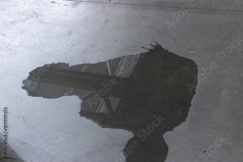 A puddle of fuel and water on a concrete floor with reflections of the ceiling and window structures inside the building