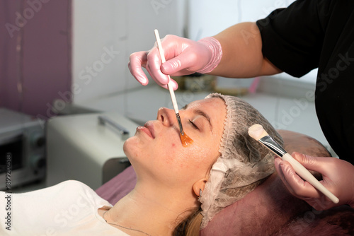 A cosmetology facial treatment is performed using a brush applicator on a woman lying on a treatment bed, with protective gloves and disposable cap visible in a professional skincare environment. Jet 