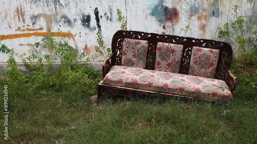 Worn wooden couch abandoned in grass near a cracked graffiti wall, symbolizing neglect, decay, and forgotten urban environments.