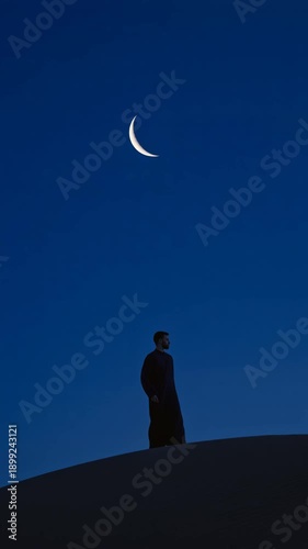 Man in traditional attire stands on a sand dune under a crescent moon in a deep blue sky