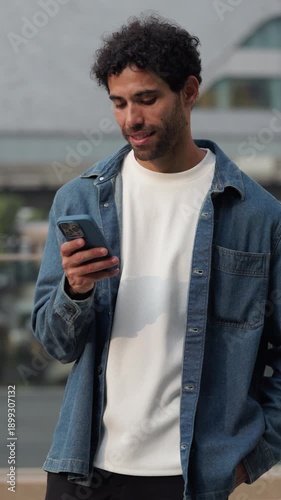 Portrait of happy Hispanic man types message on smartphone as stands in downtown vertical shot. Latin guy scrolls social media via mobile phone in summer city. Digital device