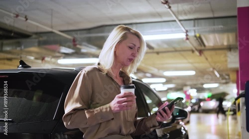 Middle aged blonde woman drinking coffee and using smartphone while waiting for electric car to charge. Concept of eco friendly transport, electric vehicle lifestyle, sustainability and urban mobility