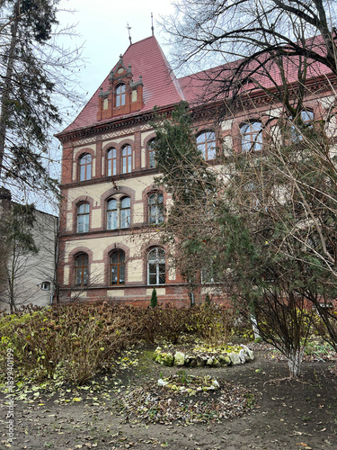 historic brick building with a steep red tiled roof and arched windows, partially framed by bare trees and shrubs in late autumn, Transcarpathia, Beregovo