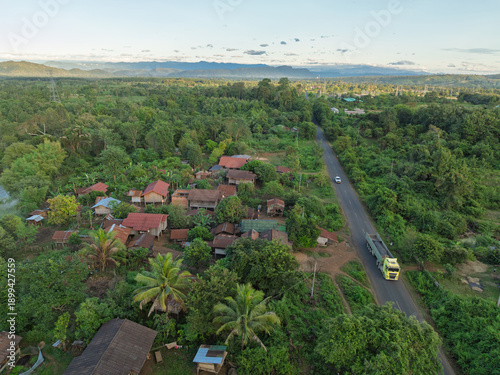 Drone view of rural settlement beside a paved road cutting through lush forested countryside in southern Laos.