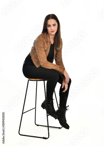 full length portrait of pretty brunette female model wearing basic modern  black clothing with brown leather jacket. sitting pose on chair, isolated figure on white studio background.
