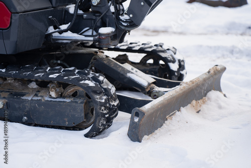 Tracked mini excavator chassis on snow with dozer blade and hydraulic parts, close up of rubber track system, idler wheels and metal plow for cold season worksite operation