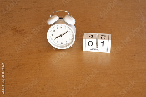 A white alarm clock sits on a wooden table next to a calendar. The clock reads 10:30 and the calendar is marked with the date April 1