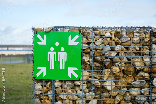 Panneau carré sur fond vert illustrant un point de rassemblement avec quatre flèches pointant deux silhouettes, accroché sur un mur de séparation, composé de roches dans un grillage