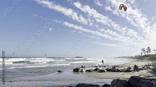 Mui Ne, Vietnam – February 2, 2026: Wide beach scene with multiple kites in the sky and surfers riding waves in strong wind conditions. Popular kitesurfing spot in Vietnam. 