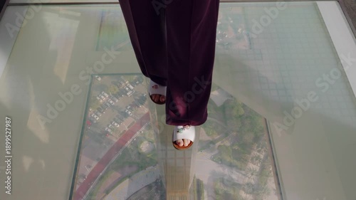 Middle aged caucasian woman walking on transparent glass floor indoors at modern observation deck, city visible below, symbolizing adventure travel and unique urban experience.